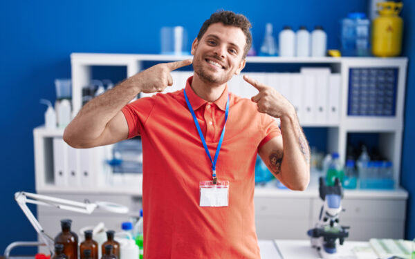 young hispanic man working at scientist laboratory smiling cheerful showing and pointing with fingers teeth and mouth. dental health concept.