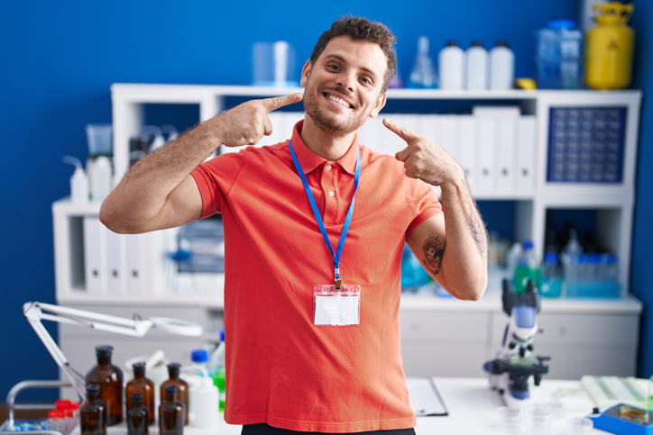 young hispanic man working at scientist laboratory smiling cheerful showing and pointing with fingers teeth and mouth. dental health concept.